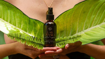 Hands holding Lab.Otanica tonic surrounded by tropical leaves symbolizing organic skincare and earth-honoring beauty rituals.